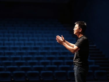 A speaker on stage, seen from the side, gesturing confidently under a warm spotlight, empty auditorium, deep blue and slate tones, high contrast, 35mm lens.