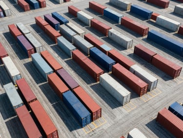 Wide-angle drone shot of an industrial logistics hub, rows of shipping containers, overhead view, crisp geometric shadows, bright daylight.