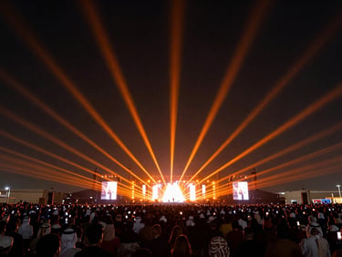 Wide-angle shot of forty orange laser beams shooting from a massive outdoor stage into a dark night sky in Dammam, massive crowd, high contrast
