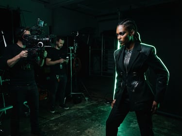 Behind-the-scenes shot of a high-fashion model posing in a dark studio with sharp neon green rim lighting, a camera and crew visible in the background, 85mm lens, high-contrast editorial fashion photography.