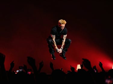 High-contrast concert photography of Falkon King jumping mid-air on a dark stage, dramatic crimson backlighting, crowd silhouettes below.
