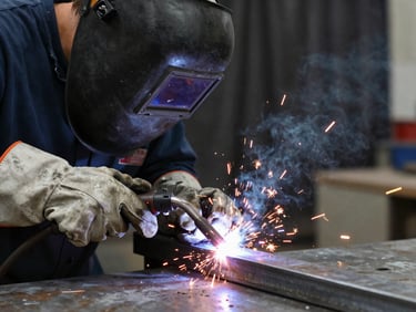 Welder's gloved hands guiding a MIG torch along a steel joint, bright arc light and orange sparks, dark welding curtain in the background, metal table surface visible