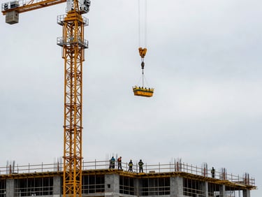 Tower crane rising above a building frame at overcast daylight, load suspended mid-air, construction workers visible at base engaging with rigging, dust and wear on equipment