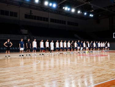 Wide shot of an indoor sports arena, clean wooden court, organized athletic teams standing in line, bright overhead lighting.