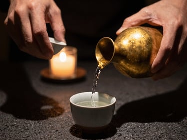Hands pouring sake from a warm brass carafe into a small ceramic cup, deep shadows, shallow depth of field, warm lighting.