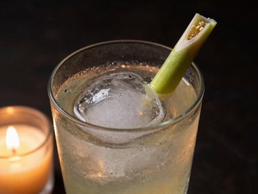 Close-up of a cocktail glass with a clear ice sphere and bruised lemongrass garnish, moody candlelight, dark background.