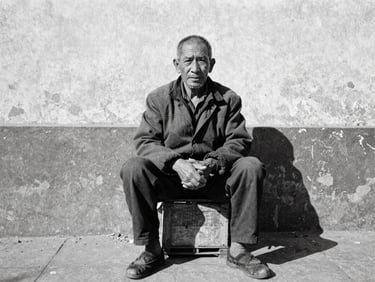 Candid environmental portrait of an elderly street vendor sitting on a wooden crate, high-contrast shadows, natural overcast daylight, gritty 35mm grain