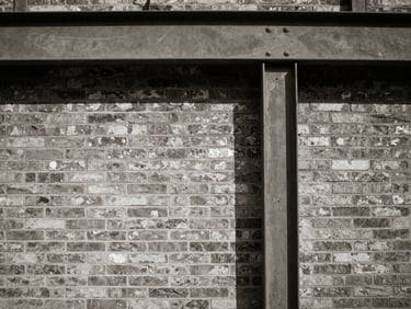 Medium-format photograph of a brick wall meeting a steel beam, high-contrast shadows, raw textures, muted tones.