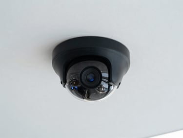 Discreet black dome security camera mounted on a clean white office lobby ceiling, minimalist architecture, soft cool lighting, sharp focus