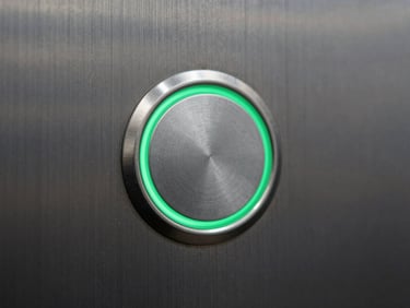 Macro close-up of a brushed stainless steel elevator button panel, with a single button highlighted by a clean, thin emerald green LED ring. Cool studio lighting.