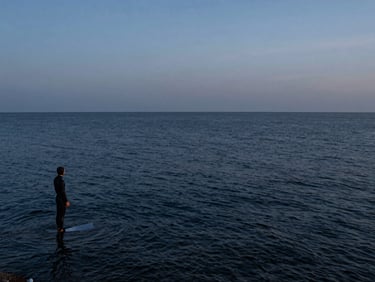 Wide panoramic pre-dawn coastline, a lone freediver in a wetsuit standing at the ocean's edge in the far left third, dark water stretching to the horizon, blue-hour ambient light, documentary-style unposed moment