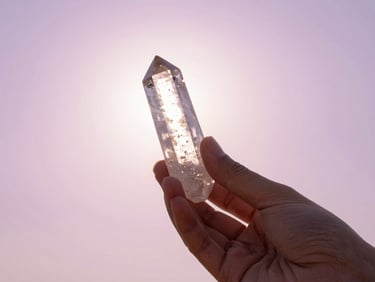 A hand holding a clear quartz crystal against a soft pastel pink and lavender sky, sun flare, dreamlike atmosphere.