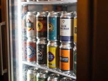 Close-up of craft beer cans with colorful labels stacked neatly behind a glass cooler door with condensation