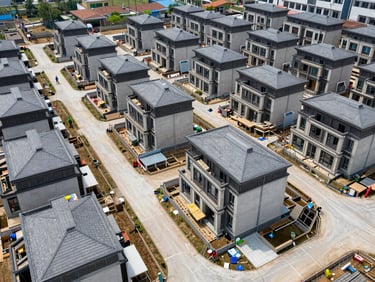 Overhead site photo of a residential compound under final finishing, natural daylight, rows of completed villas visible with paved internal roads, workers in hard hats in the distance
