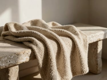 Undyed oatmeal wool blanket draped softly over a rustic plaster bench, casting long warm shadows
