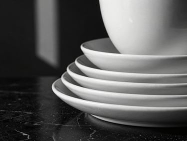 Extreme close-up of premium matte ceramic tableware stacked on a polished black marble surface, directional window light, deep shadows
