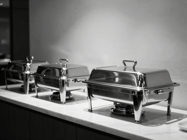 Minimalist buffet setup in a luxury hotel atrium, polished glass and steel chafing dishes, bright highlights and deep shadows.