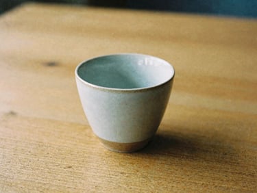 A single ceramic cup resting on a textured wooden table, soft shadows, warm earth tones, calm atmosphere, 35mm film aesthetic