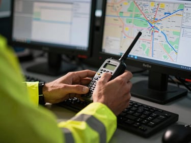 Close-up of a dispatcher's hands on a radio and computer screen, with a logistics map visible, in the Red Oak office, high-visibility gear, hard daylight, industrial editorial photography, sharp details