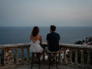 A quiet moment of a couple on a stone balcony overlooking the Mediterranean at dusk, moody overcast natural light, analog film grain, documentary style.
