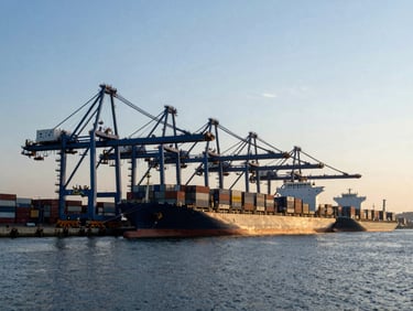 Wide environmental shot of a deep-water shipping port at dawn, massive cargo cranes silhouetted against cool morning daylight, container vessel docked.