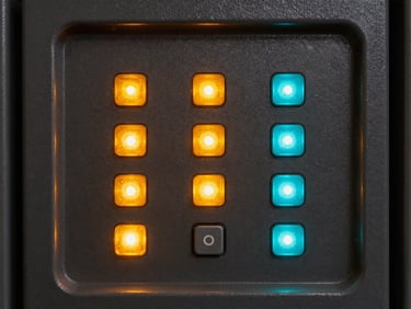 Close-up of an alarm control panel with illuminated status LEDs in amber and cyan, keypad buttons in sharp focus, dark metal housing with rim lighting revealing form detail