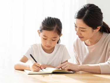 A mother helping her daughter write in a notebook at a sunlit wooden table, bright high-key lighting, soft clean colors