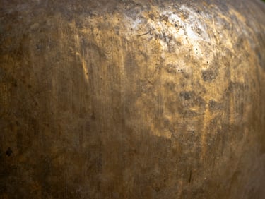 Extreme close-up 16:9 horizontal crop of a patinated bronze surface, hand-worked oxidation marks visible across the grain, studio tungsten light raking at a low angle to reveal surface topography, no context objects, purely the metal finish in transition