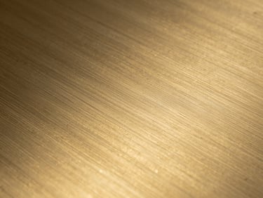 Extreme tight crop of a brushed brass surface, grain direction visible under studio tungsten raking light, hand-finishing marks present, 16:9 horizontal, purely the metal surface