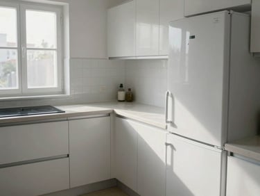 Wide shot of a furnished studio kitchen corner, open-plan counter with induction hob and under-counter fridge, white tiles, morning window light casting clean shadows on the counter surface