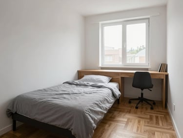 Eye-level wide shot of a compact furnished studio, single bed with grey linen against a white wall, wooden desk under a window flooding the room with morning daylight, parquet floor visible