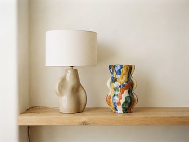 Vue latérale d'une lampe sculpturale en céramique beige et d'un vase coloré, posés sur une étagère en bois clair, lumière naturelle diffuse, arrière-plan minimaliste crème, profondeur de champ faible, 35mm.