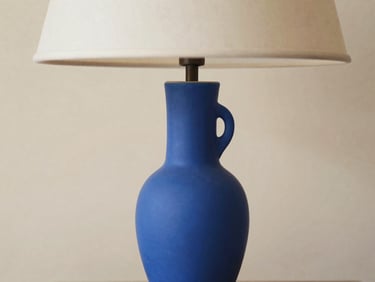 Close-up portrait photograph of a unique, sculptural ceramic table lamp in vibrant blue, minimalist composition on a textured beige background, soft diffused daylight, shallow depth of field, 35mm.