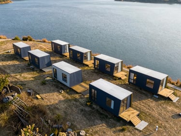 Aerial drone shot of a modern glamping resort with five modular tiny houses arranged neatly along a lake, golden hour lighting