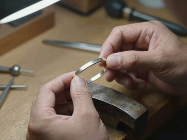 Close-up of jeweler's hands holding silver bracelet under diffused studio light, workbench surface visible in background with small tools, warm honest workshop atmosphere, shallow depth of field