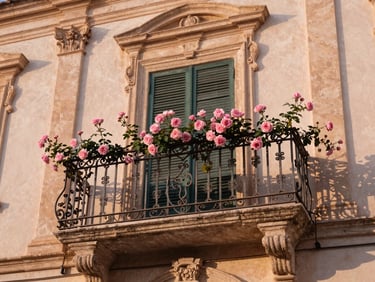 Warm editorial shot of a baroque Sicilian balcony with delicate ironwork, soft pink climbing roses, warm afternoon light.