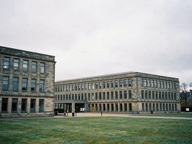 Cool editorial photography, overcast northern daylight, desaturated slate tones, sharp focus, 35mm film. A wide environmental view of stone-built offices at High Fernley Court in Wyke, Bradford, showing clean industrial architecture and a grey sky.