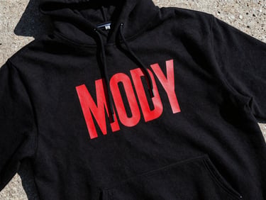 Moody high-contrast close-up of a bold graphic print on a heavy black cotton hoodie, harsh direct sunlight, stark shadows, raw concrete background, 35mm