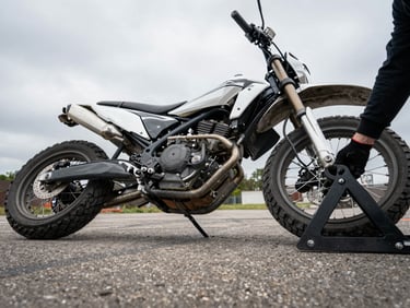 Low-angle shot of a fallen motorcycle on pavement, a gloved hand gripping a lift frame positioned under the subframe, overcast daylight, no bystanders, industrial recovery framing