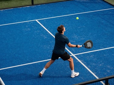 Close overhead angle of a single player mid-backhand rally on a padel court, natural daylight, court lines and blue surface texture sharp, player in focused athletic motion, no posed expression