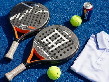 Overhead close-up of premium padel rackets, a can of balls, and a folded polo shirt arranged on a court-blue surface under sharp natural daylight, clean geometric shadow lines across the gear
