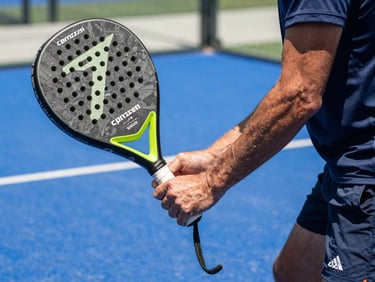 Close-up of a coach demonstrating forehand grip on a padel racket, court surface visible in background, sharp natural daylight, focused and authoritative framing