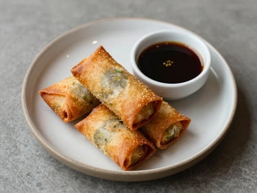 Overhead shot of a plated starter — crispy golden spring rolls and a small dipping sauce on a ceramic plate, natural daylight, minimal styling