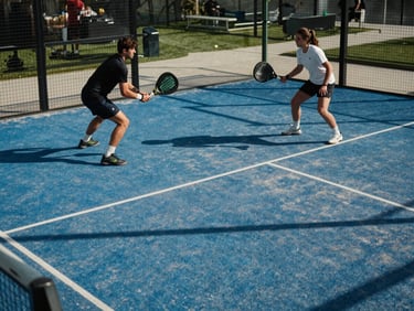 Wide shot of a padel court mid-rally, two players in motion, sharp natural daylight casting crisp court-line shadows on the blue-green playing surface, decisive action framing