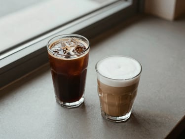 Overhead shot of an iced coffee and a frappuccino in clear glasses on a cafe counter, ice visible, natural side-window light, minimal styling