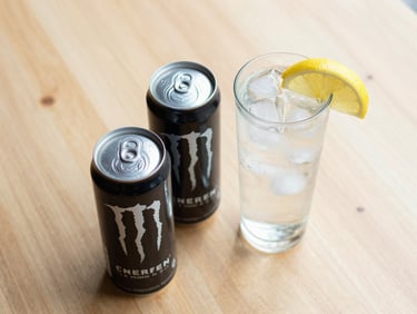 Overhead shot of two cans of energy drinks beside a tall glass of cold drink with ice and a lemon slice on a light wooden surface, natural daylight, clean minimal background