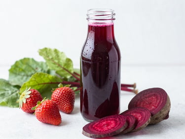 A glass bottle filled with deep red beet juice, next to fresh strawberries and sliced beetroot on a high-key light surface