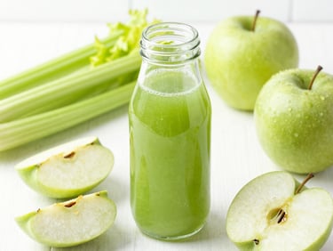 A glass bottle filled with vibrant green juice, surrounded by fresh sliced green apples and celery stalks on a bright white surface, natural lighting