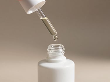 A white ceramic dropper bottle with a single droplet of clear oil hanging from the pipette, soft shadows