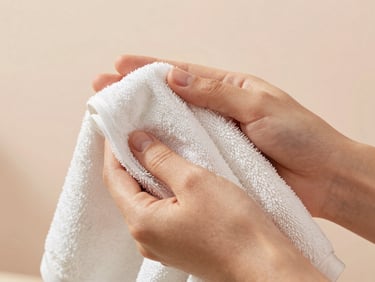 Close-up of gentle hands holding a warm damp organic cotton towel, soft ambient light, warm blush background, editorial skincare photography